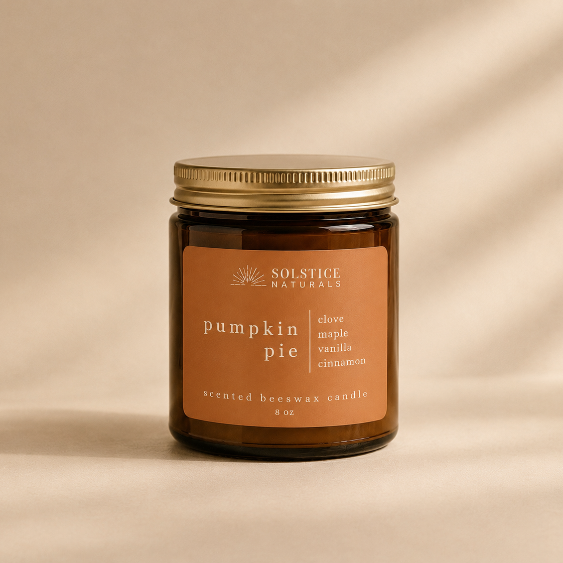 A single amber glass jar candle with a gold metal lid and a warm tan label reading 'SOLSTICE NATURALS pumpkin pie' on a softly lit beige background.